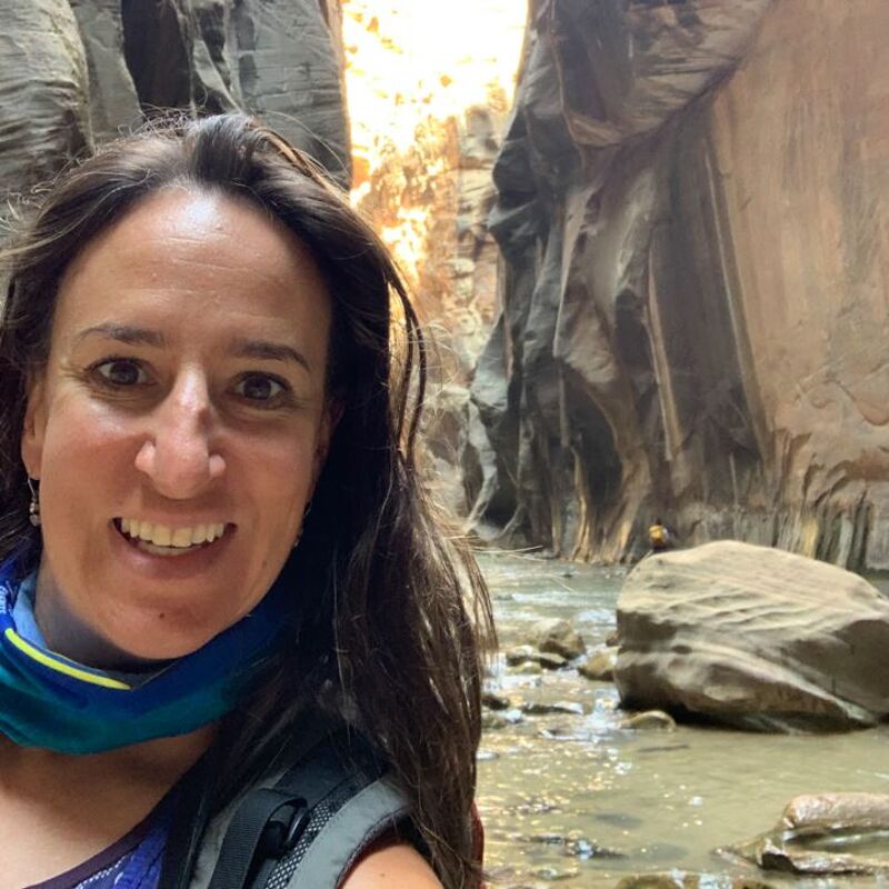 The image shows a woman in a canyon with a stream. She is smiling at the camera. The canyon walls are tall and made of smooth rock. The stream is shallow and rocky. The woman is wearing a backpack and a blue and yellow scarf. It looks like she is hiking or exploring the canyon.